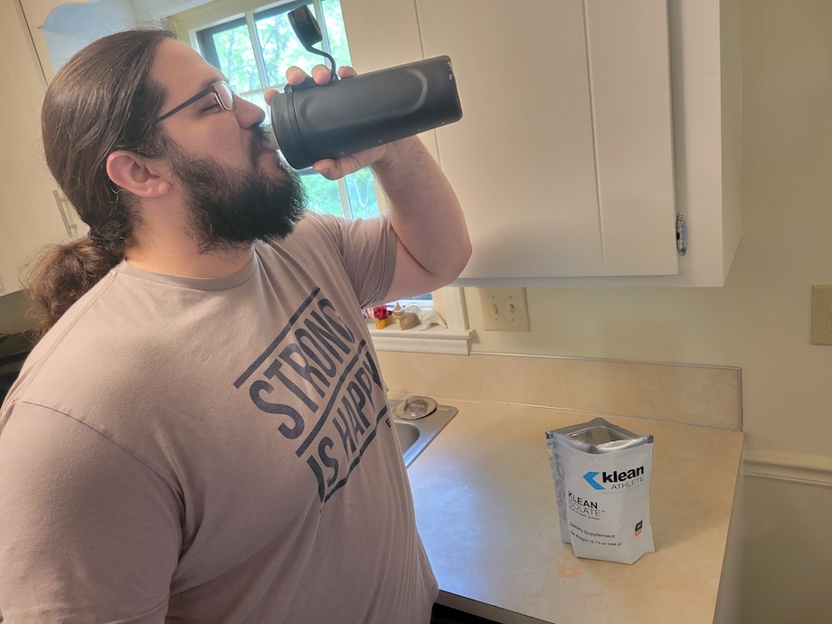 A person with long hair and a beard, wearing a Strong is Happy shirt, drinks from a black bottle in a kitchen. A packet labeled Klean is on the counter beside them.