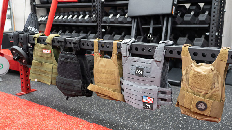 Five weighted vests and one weight plate carrier hanging side-by-side for comparison on a metal bar, sitting in front of a dumbbell rack.