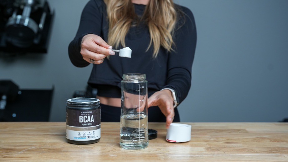 A woman taking a scoop of Jacked Factory Fermented BCAAs and placing them in a shaker cup filled halfway with water