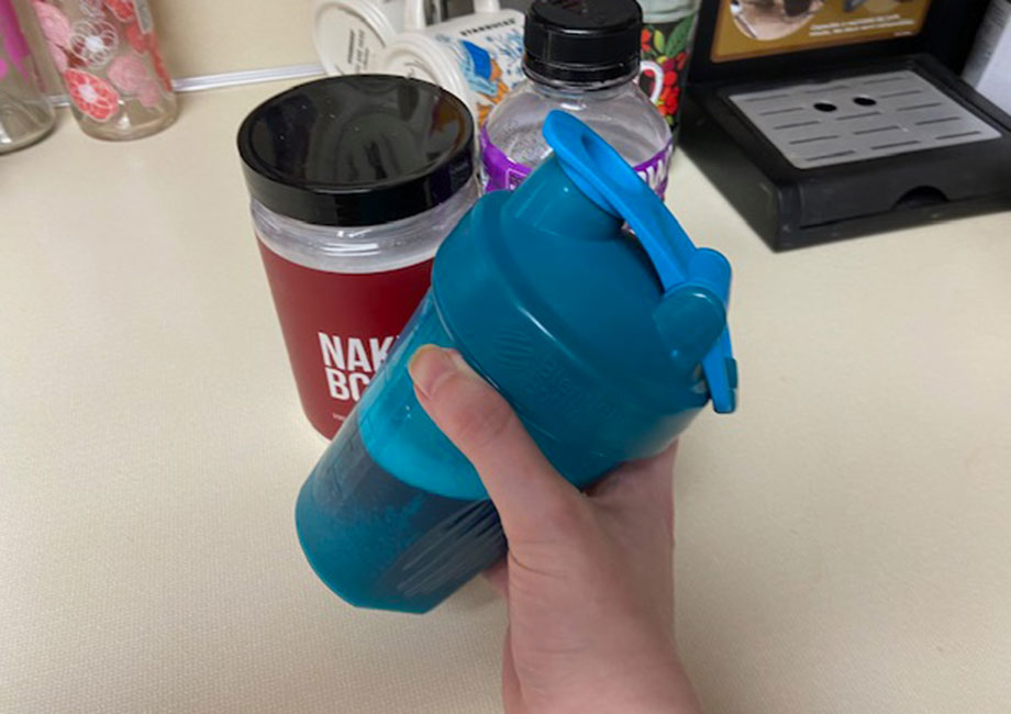 A hand holding a blue shaker bottle in front of a kitchen counter. Nearby are a red container labeled NAK and purple and black bottles. A coffee pod holder is visible in the background.