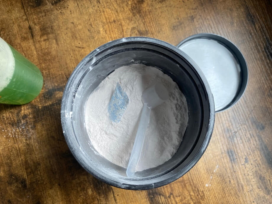 Open container of white powder with a plastic scoop inside, sitting on a wooden surface. A green shaker bottle is partially visible on the left.