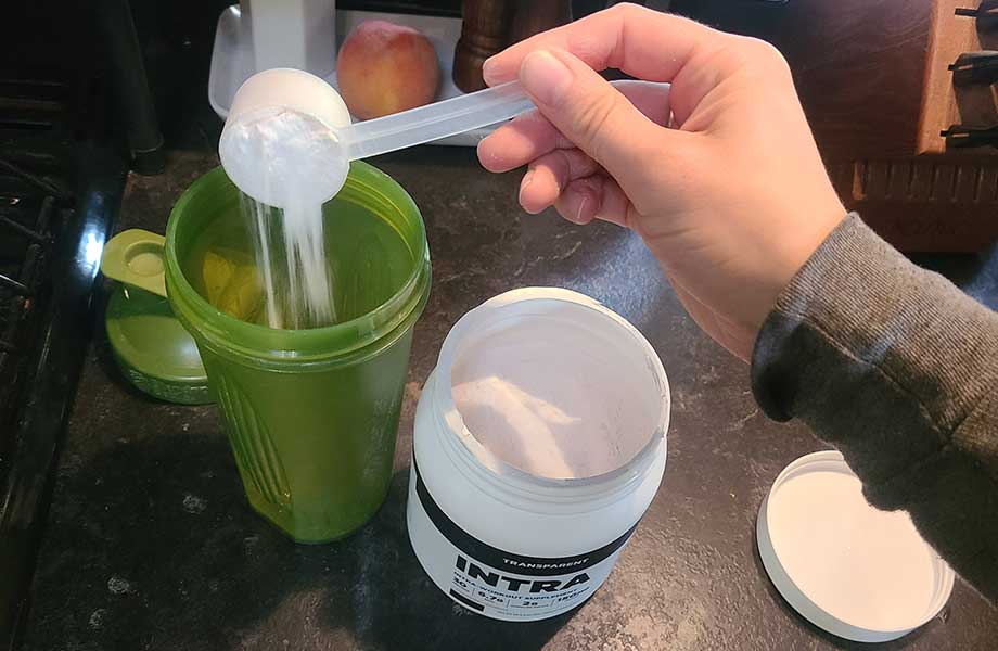 A hand is scooping white powder from a container labeled INTRA into a green shaker cup on a kitchen counter. A peach and other kitchen items are visible in the background.