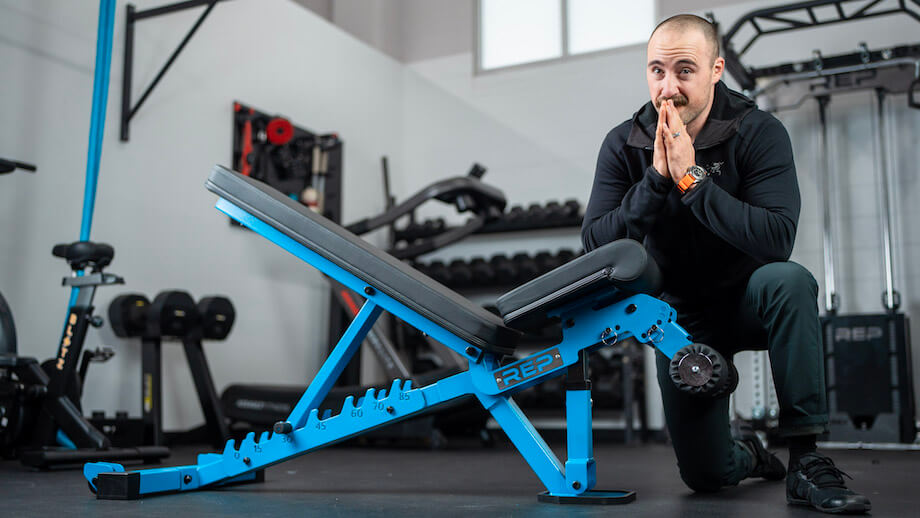 Coop kneels next to a REP Fitness AB-3000 2 weight bench. The bench is blue with a black pad.