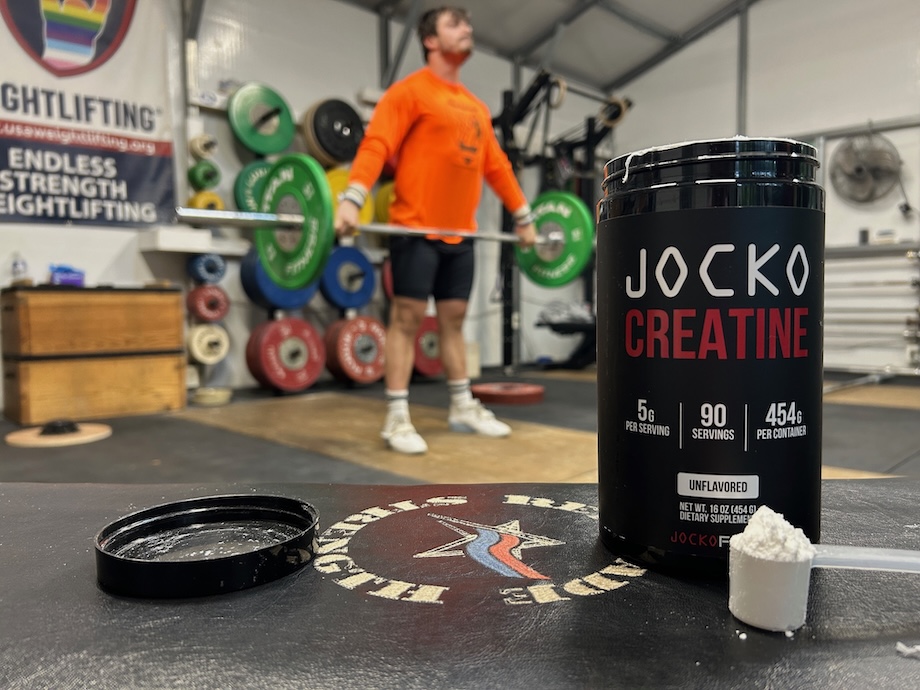 Container of Jocko Creatine on the floor of a gym in front of a man performing Olympic lifts.