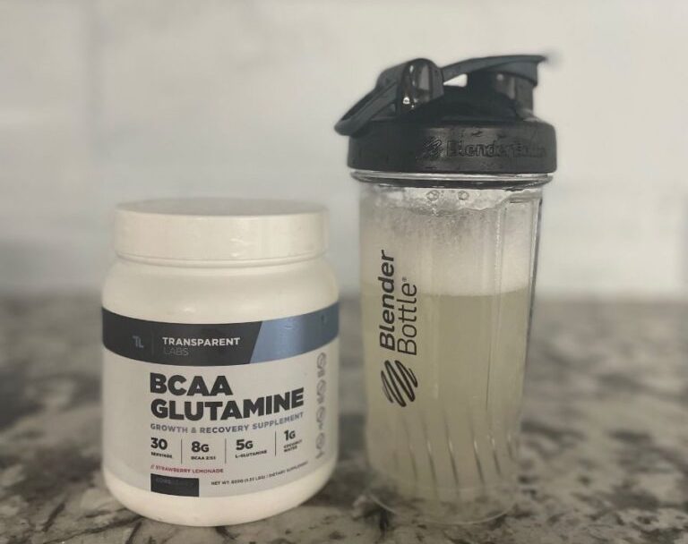A tub of BCAA Glutamine supplement is next to a Blender Bottle filled with a light-colored liquid, placed on a marble countertop. The BCAA label highlights 30 servings, 8g BCAA, and 5g Glutamine.