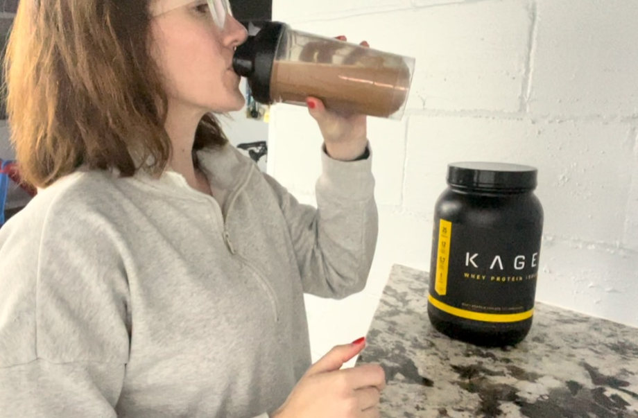 A woman drinking chocolate Kaged Whey Protein Isolate out of a clear shaker bottle with the container of protein powder in the background